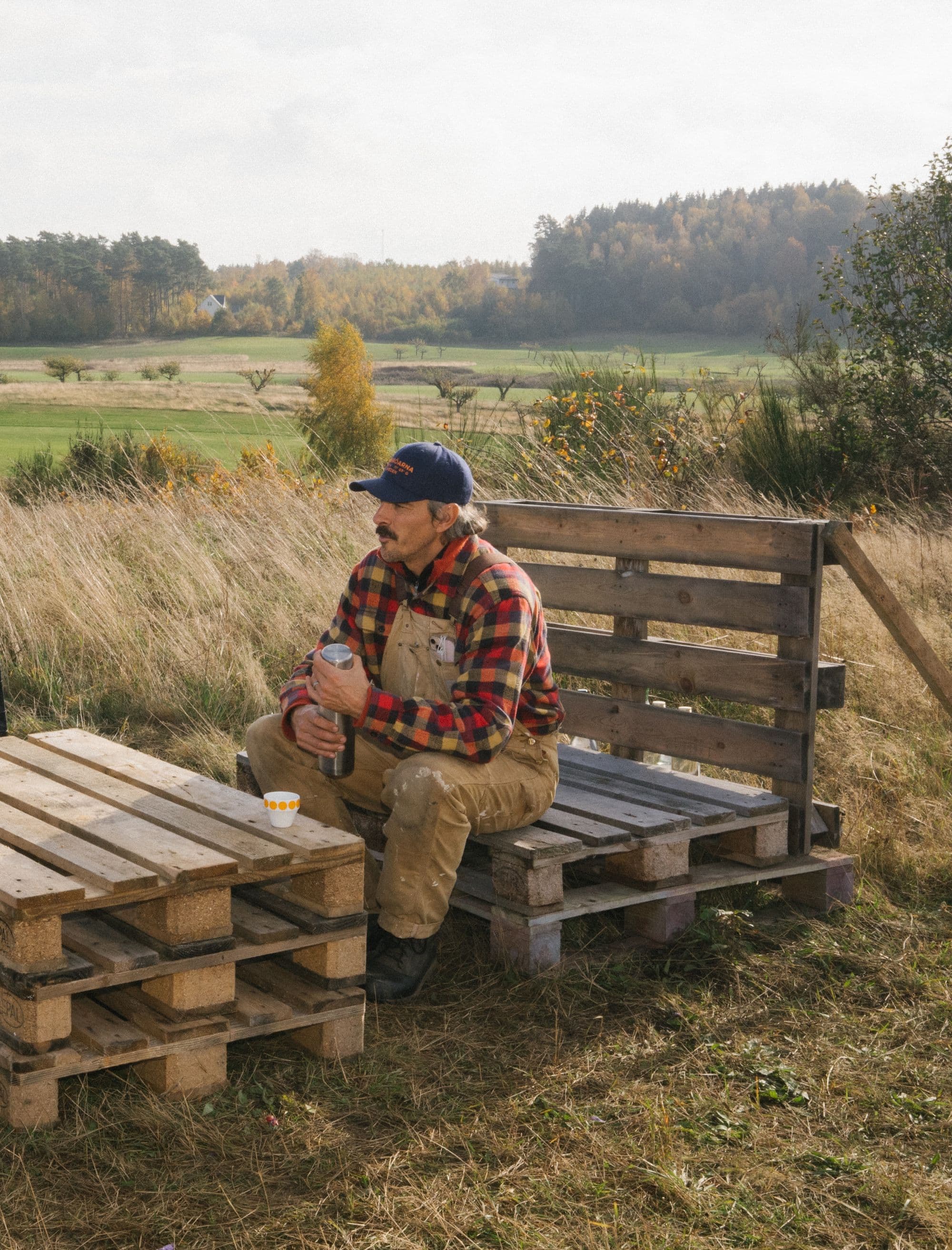 A man in a plaid shirt and overalls sits on a wooden pallet bench, holding a thermos, with a pallet table and cup nearby in a field with autumn trees in the background.